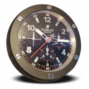 White background with a circular wall clock that is in the style of a HUBLOT Big Bang wristwatch. It has a black bezel with six signature HUBLOT screws around it in the two, four, six, eight, ten and twelve o'clock positions. The dial is all black with a chequered flag type pattern to it and red second indices that span around the very outside. There are main numbers and indices on the dial that stand prominent and are a brushed silver colour. On the lower part of the dial is three sub dials, each with silver colour hands and more red indices. A white HUBLOT 'H' logo and text reading 'HUBLOT' then 'GENEVE' written underneath each other, sit just under the twelve o'clock position at the top middle of the dial. There is a long red second hand with a HUBLOT 'H' on the shorter end of it. The minute and hour hands are a polished silver colour with luminescent (Glow in the dark) centres.