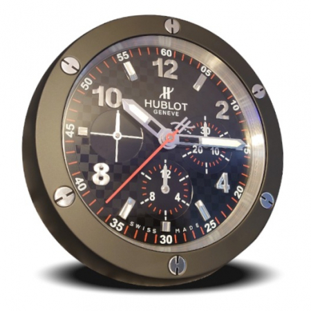 White background with a circular wall clock that is in the style of a HUBLOT Big Bang wristwatch. It has a black bezel with six signature HUBLOT screws around it in the two, four, six, eight, ten and twelve o'clock positions. The dial is all black with a chequered flag type pattern to it and red second indices that span around the very outside. There are main numbers and indices on the dial that stand prominent and are a brushed silver colour. On the lower part of the dial is three sub dials, each with silver colour hands and more red indices. A white HUBLOT 'H' logo and text reading 'HUBLOT' then 'GENEVE' written underneath each other, sit just under the twelve o'clock position at the top middle of the dial. There is a long red second hand with a HUBLOT 'H' on the shorter end of it. The minute and hour hands are a polished silver colour with luminescent (Glow in the dark) centres.